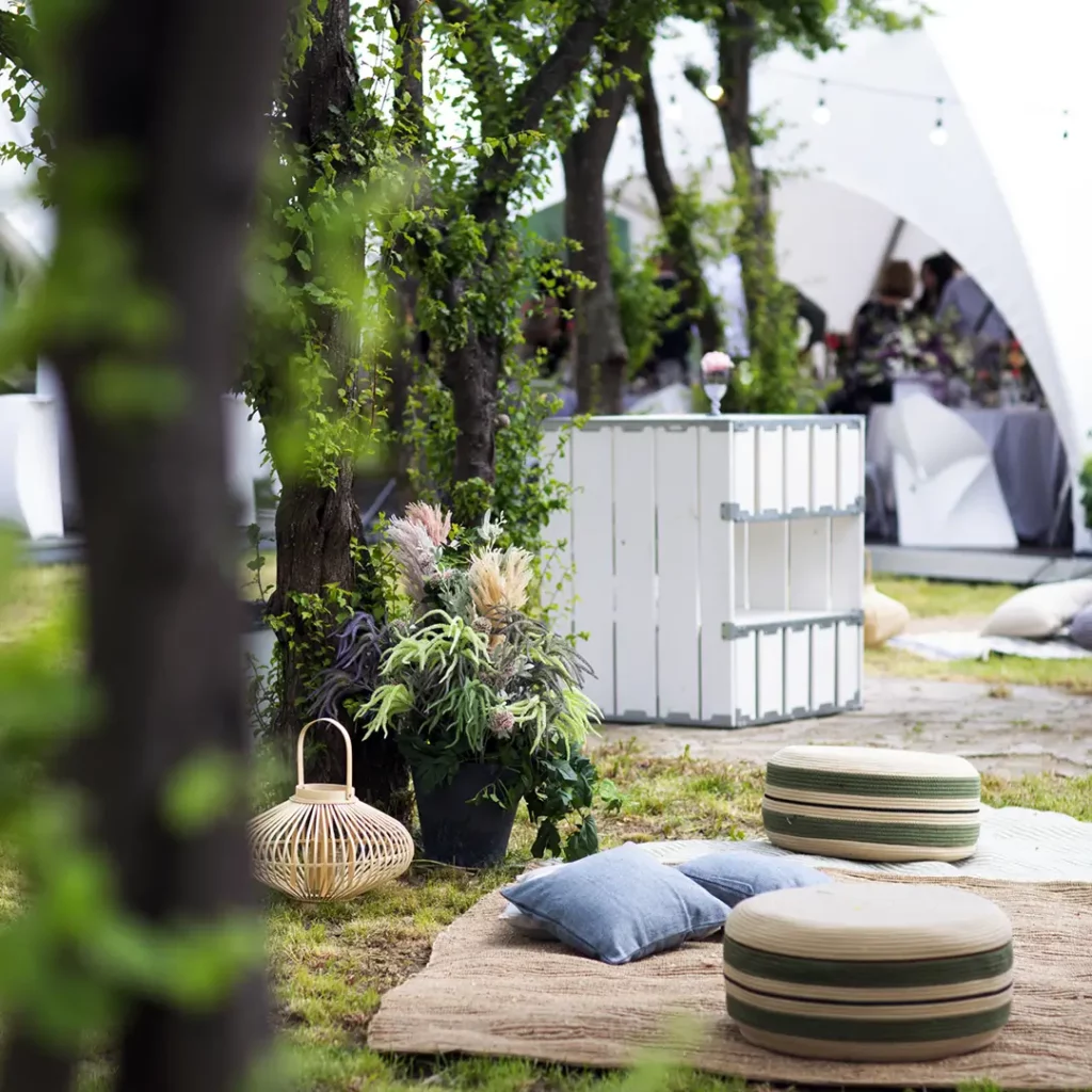 Location de chapiteau : Tentation et ses partenaires déco créent une ambiance féérique pour une cérémonie au parc. En premier plan, un espace cosy avec tapis, coussins, et poufs, accompagnés d'un panier en osier design. En second plan, des mange-debout en bois blanc et vert alignés dans la profondeur. En arrière-plan, une scène de cérémonie abritée par un chapiteau dôme Tent'Action.