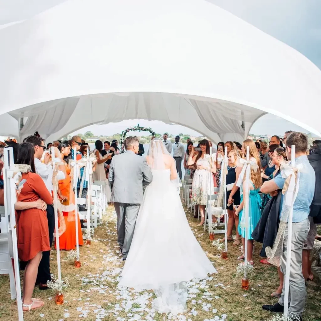 Organisation Événementielle 360, Scène de mariage : Un couple de mariés avance vers l'autel pour la cérémonie, abrité par un dôme Tent'Action au bord d'un lac. L'ensemble des proches est réuni pour célébrer ce moment spécial.